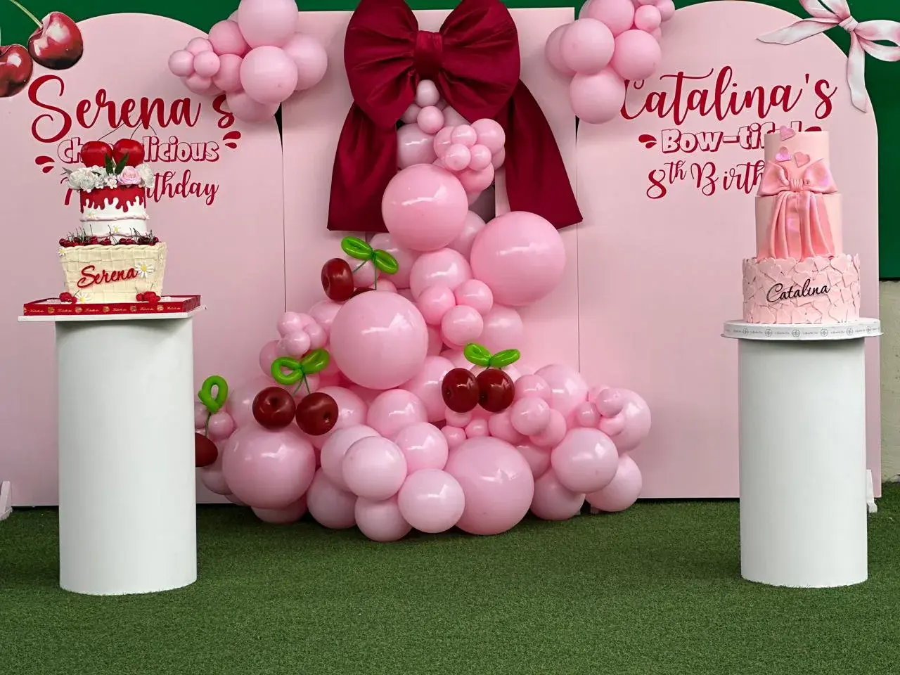 Pink birthday setup with balloons, a big bow and two cakes on the turf